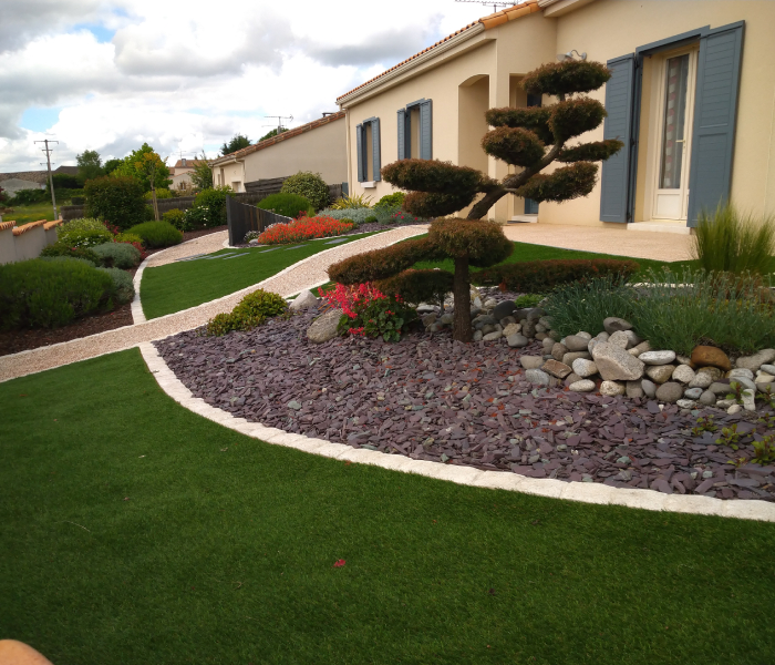 Réalisation paysagère avec pelouse, massif décoratif en pierre et arbustes structurés autour d’une maison